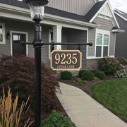 A scalloped cast bronze aluminum address plaque hanging from a decorative address plaque post in front of a house