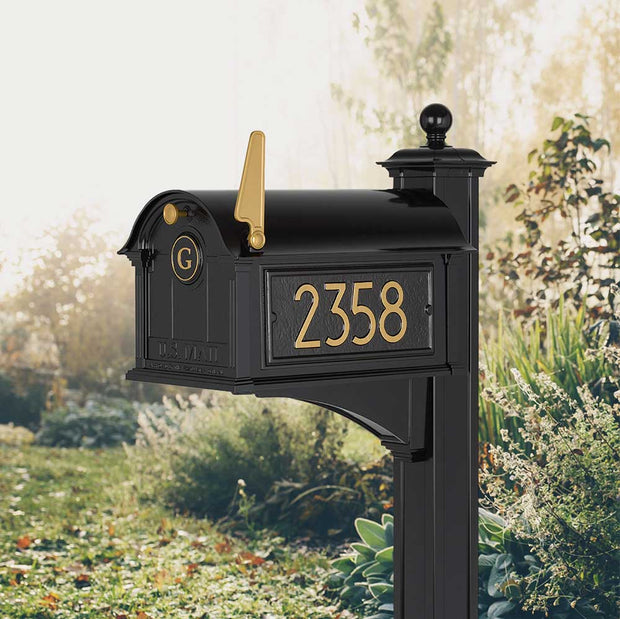 Modern Balmoral Mailbox shown in front of green foliage. Mailbox shown with a black finish and gold accents. 