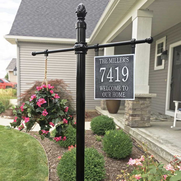 An example square address sign post assembly with a post-top light, a hanging flower basket on one side, and the hanging address plaque on the other side, all in front of a grey house